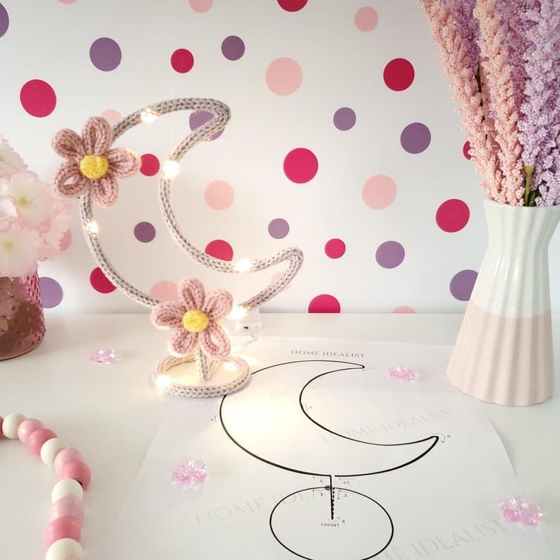 May include: A handcrafted, gray crescent moon-shaped lamp with embedded lights and two pink crochet flowers with yellow centers. The lamp sits on a white surface with a floral pattern and a paper with a moon outline.