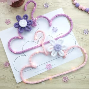 May include: Two handmade, heart-shaped crochet hangers in light pink and lavender, adorned with crocheted flowers. The hangers are embellished with tiny lights, and sit on a white paper with a cloud outline. Decorative floral accents are scattered around.