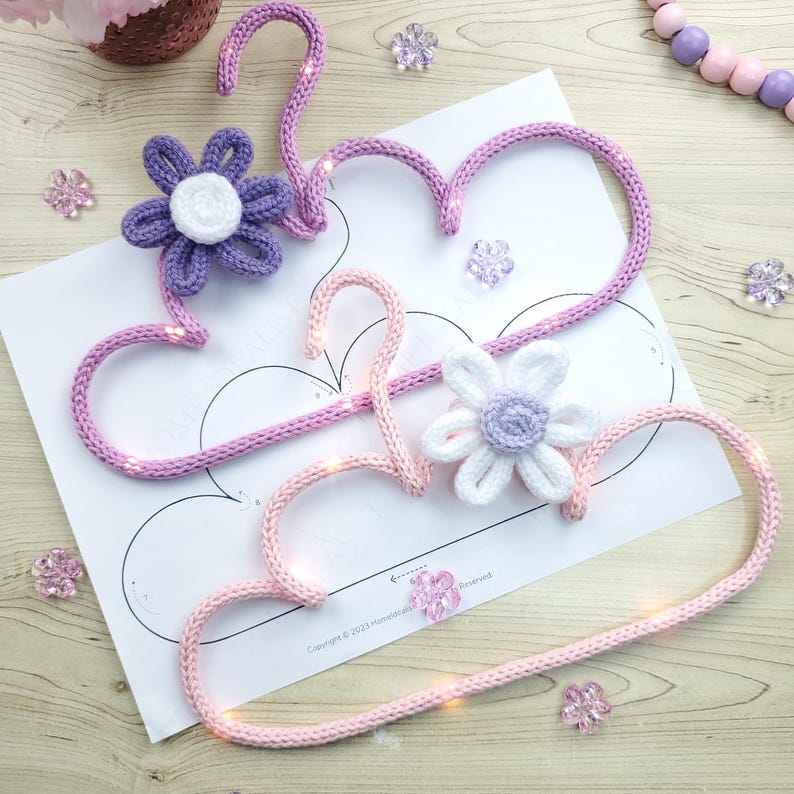 May include: Two handmade, heart-shaped crochet hangers in light pink and lavender, adorned with crocheted flowers. The hangers are embellished with tiny lights, and sit on a white paper with a cloud outline. Decorative floral accents are scattered around.