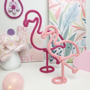 May include: Two knitted flamingo sculptures in shades of pink and fuchsia stand on a white surface. The flamingo figures are made of thick yarn and have a whimsical, decorative design. A knitted flower and a decorative ball are also visible.