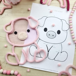 May include: A pink knitted pig-shaped item with black eyes and a beige snout sits next to a pig illustration on a white paper. The paper has the text "HOME IDEALIST" and "Copyright &copy; 2024 Homeldealist". Pink pom-poms and a beaded garland are also present.