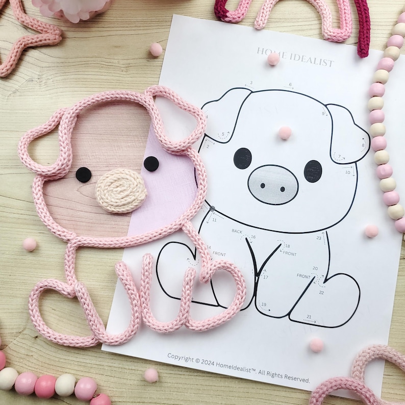 May include: A pink knitted pig-shaped item with black eyes and a beige snout sits next to a pig illustration on a white paper. The paper has the text "HOME IDEALIST" and "Copyright &copy; 2024 Homeldealist". Pink pom-poms and a beaded garland are also present.