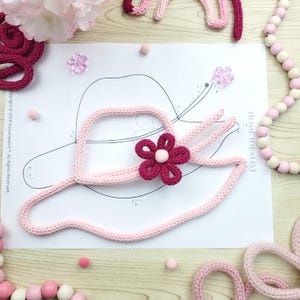 May include: A light pink knitted hat with a dark pink flower and a white center, placed on a paper with a hat outline. The image includes pink pom-poms, flower-shaped embellishments, and a pink and white beaded garland. The text "HOME IDEALIST" is visible.