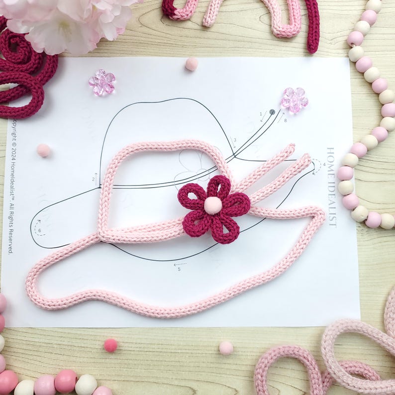 May include: A light pink knitted hat with a dark pink flower and a white center, placed on a paper with a hat outline. The image includes pink pom-poms, flower-shaped embellishments, and a pink and white beaded garland. The text "HOME IDEALIST" is visible.