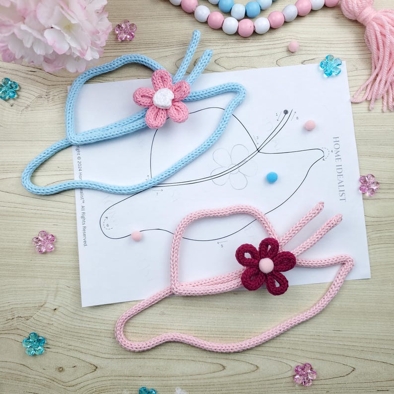 May include: Two handmade, knitted sun hats with flower accents. One hat is light blue with a pink and white flower, the other is pink with a burgundy flower. The hats are placed on a paper with a hat outline and the text "HOME IDEALIST".