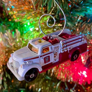 May include: A white and red fire truck Christmas ornament. The ornament has a silver wire hanger and features the text "UNIT 12". The fire truck has black wheels and red accents.