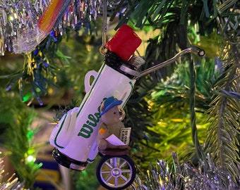May include: A white golf bag ornament with a green and white "Wilson" logo. A small brown bear figurine is sitting in the bag, holding a sign that says "Score Card".