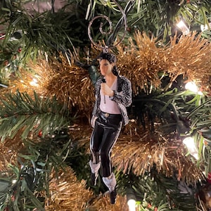 May include: A Christmas tree ornament depicting a figure in a black sequined jacket, white shirt, and black pants, holding a microphone. The ornament has a silver metal hanger and is set against a backdrop of green and gold Christmas tree decorations.