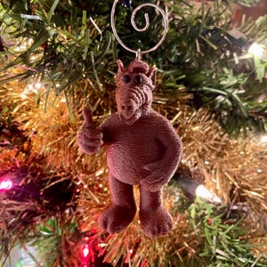May include: A brown, 3D-printed Christmas tree ornament shaped like the character Alf, giving a thumbs-up gesture. The ornament is suspended by a silver wire and hangs against a backdrop of a decorated Christmas tree.