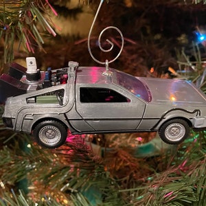 May include: A silver-colored DeLorean time machine ornament hanging from a Christmas tree. The ornament is detailed with a black interior and a silver-colored body.