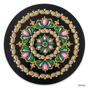 May include: A circular mandala painted on a black background with gold, pink, and green accents. The mandala features a repeating pattern of flowers and leaves.