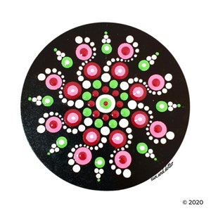 May include: A black circular mandala with a white, pink, and green dot pattern. The mandala is painted on a black background and has a swirling design.