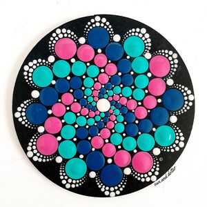 Op de afbeelding: Een cirkelvormige mandala geschilderd op een zwarte achtergrond met witte, roze, turquoise en donkerblauwe stippen. De stippen zijn in een spiraalvorm gerangschikt, waardoor een bloemvormig ontwerp ontstaat.