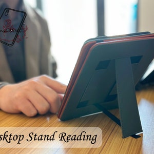 Peut inclure: Un support de tablette en cuir noir avec un accent rouge. Le support est pli&eacute; et pos&eacute; sur une surface en bois. Le texte "Desktop Stand Reading" est visible en bas de l'image.