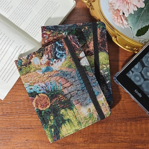 May include: Notebooks with colorful tapestry covers featuring a landscape design. The notebooks have black elastic bands and are stacked on a wooden surface. A Kindle e-reader and a book are also visible.