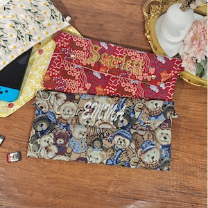 May include: Two rectangular fabric pouches with envelope-style flaps and gold-tone snap closures. The top pouch is red with a floral pattern and the name Scarlett embroidered in gold. The bottom pouch features a teddy bear print and the name Emma.