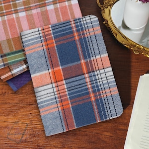 Könnte beinhalten: Notizbücher mit Karomustern in verschiedenen Farben. Das Hauptnotizbuch hat ein blaues, orangefarbenes und weißes Karomuster. Andere Notizbücher zeigen rosa, braune und violette Karomuster. Die Notizbücher liegen auf einer Holzoberfläche.