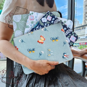 Op de afbeelding: Een set van vier geborduurde telefoonhoesjes van stof met bloemen- en vlindermotieven. De hoesjes zijn in tinten blauw, groen en wit.