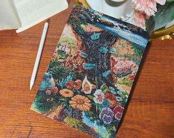 May include: A colorful tablet case with a floral and landscape design. The case features a vibrant tapestry-like pattern with flowers, trees, and a waterfall. A white stylus and an open book are also visible.