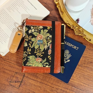 Könnte beinhalten: Ein braunes Lederjournal mit floralem Wandteppichdesign, einem schwarzen Gummiband und einem goldenen Stift. Ein Reisepass und ein Leder-Schlüsselanhänger sind ebenfalls sichtbar. Das Journal ist auf einer Textseite geöffnet.