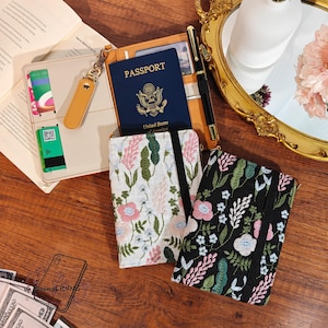 May include: An assortment of travel accessories including a passport, a tan leather wallet, a black pen, and two floral embroidered notebooks. One notebook is white with pink flowers, and the other is black with colorful flowers.