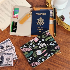 May include: An open beige and brown passport holder with a US passport, a black pen, and a black floral embroidered pouch. The pouch features pink, white, and blue flowers. US currency is visible.