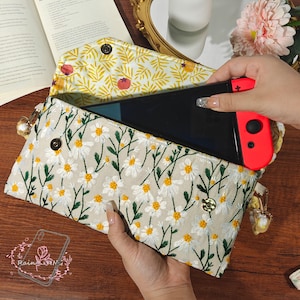 Può includere: Pochette ricamata color crema con motivo a margherite, contenente una console di gioco nera e rossa. La fodera interna presenta una stampa floreale gialla. La pochette è aperta, rivelando la console.