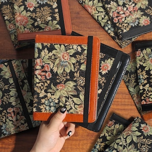 May include: Notebooks with floral tapestry covers in black, green, and red. The notebooks have brown leather trim and a black elastic band closure. Several notebooks are visible, some open and some closed.