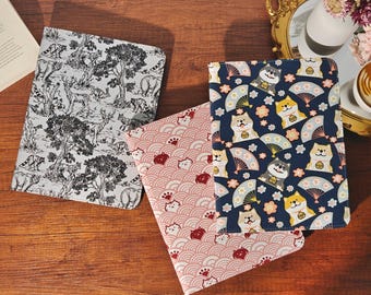 May include: Three rectangular notebooks with distinct covers. One features a black and white forest scene with animals. Another has a pink wave pattern with small cat faces. The third is dark blue with Shiba Inu dogs and fans.