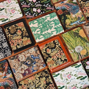 以下が含まれることがあります： 花柄、緑の松の木、テディベア、タンポポの種、風景画など、多様な模様が施された長方形の布製カバーのコレクション。各カバーには黒または茶色の無地の縁取りが施されています。