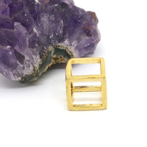 May include: A gold-colored metal cube pendant with a hollow center. The cube is made of wire and has a geometric design.