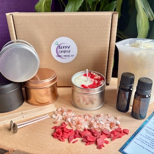 May include: A candle making kit featuring a brown cardboard box with a Berry Crafty label, three metal tins in black, copper, and silver, a plastic pouring pitcher, two amber glass bottles, wooden stir sticks, wicks, and red and pink wax bits.