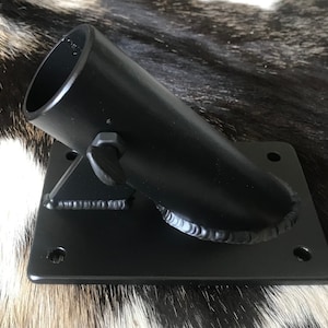 May include: Black metal flag pole holder with a square base and angled cylindrical tube. The holder has a welded construction and includes mounting holes for secure attachment. The design is simple and functional.