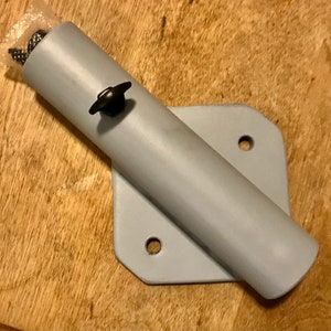 May include: A gray metal bracket with a round pipe attached. The bracket has two holes for mounting and a black knob on the pipe.