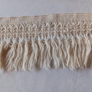 Op de afbeelding: Crèmekleurige franjeband met een geweven bovendeel en een gedetailleerd bloemmotief. De band heeft lange, vloeiende franjes, die textuur en een decoratief element toevoegen. Ideaal voor knutsel- en naaiprojecten.