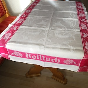 Pode incluir: Uma toalha de mesa de linho branco com uma borda vermelha e um padrão floral. A toalha de mesa tem a palavra "Rolltuch" bordada em vermelho na borda.