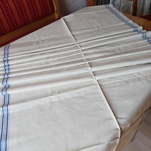 Pode incluir: Uma toalha de mesa dobrada, de cor branco sujo, com listras azuis nas bordas. O tecido é cuidadosamente plissado, criando uma superfície texturizada. A toalha de mesa é provavelmente feita de um material natural e durável.