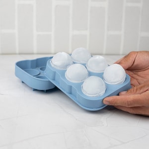 Peut inclure: Un bac à glaçons en silicone bleu avec six glaçons ronds. Le bac est ouvert et les glaçons sont visibles.