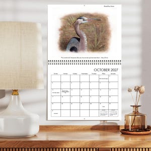 May include: A wall calendar for October 2027 featuring a Great Blue Heron illustration at the top. The page includes the quote, "This world is full of poems. But you have to be quiet to hear them. - Mary Oliver," and a standard grid for the month.