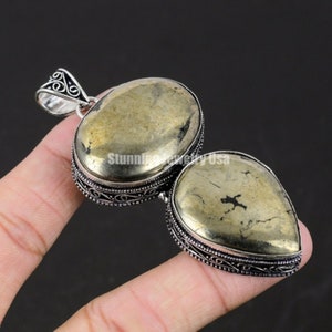 Puede incluir: Un colgante de plata con dos piedras de pirita en forma de lágrima. Las piedras son de color dorado con motas negras. El colgante tiene un intrincado diseño de plata alrededor de los bordes.