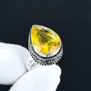 May include: A silver ring featuring a large, pear-shaped yellow gemstone. The gemstone is faceted, catching the light and displaying a vibrant color. The ring has an ornate, detailed setting with a decorative border.