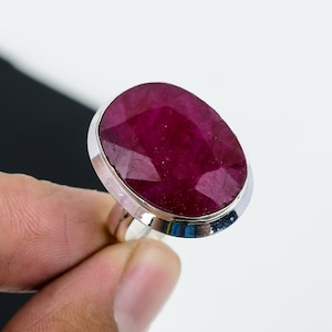 May include: A silver ring featuring a large, oval-cut ruby gemstone. The ruby is a deep, rich red color and is set in a polished silver band. The gemstone has multiple facets, reflecting light. The ring is a statement piece.