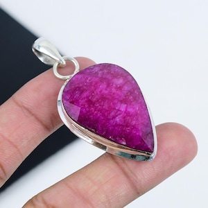 May include: A teardrop-shaped pendant with a vibrant magenta gemstone set in a silver-colored metal frame. The gemstone has a textured appearance and is suspended from a silver bail. The pendant is held by a hand.