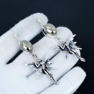 Puede incluir: Un par de pendientes colgantes plateados. Cada pendiente presenta una piedra ovalada pulida sobre un colgante de hada detallado con alas extendidas. Los pendientes se muestran sobre un guante blanco, con un fondo oscuro.