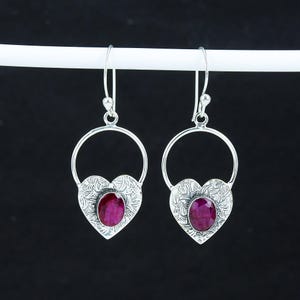 May include: A pair of silver earrings with a heart-shaped design. Each earring features a red gemstone set in the centre of the heart.