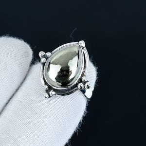 Puede incluir: Un anillo de plata con una piedra iridiscente grande en forma de pera. La piedra está engastada en una banda de plata detallada con pequeños detalles decorativos de plata. El anillo se muestra sobre un fondo blanco.