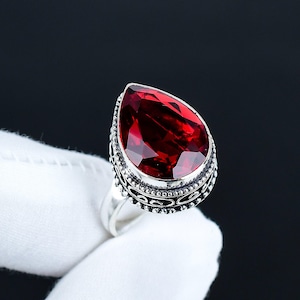 Könnte beinhalten: Ein Silberring mit einem großen, facettierten, tropfenförmigen, tiefroten Edelstein. Der Edelstein ist in einer detaillierten, verzierten Lünette gefasst. Der Ring hat ein schlichtes, poliertes Silberband.