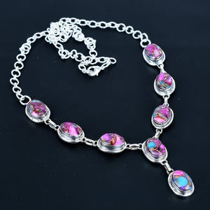May include: A silver necklace featuring oval and teardrop-shaped stones in shades of pink, purple, and blue. The stones are set in silver bezels and connected by a delicate silver chain. The necklace is displayed against a black background.
