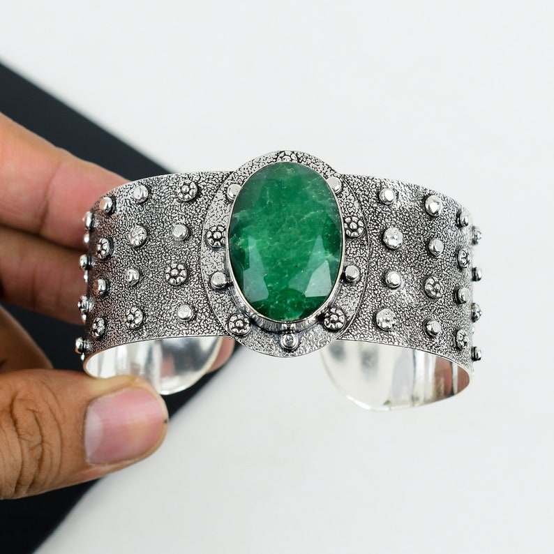 May include: A silver cuff bracelet with a large, oval-shaped green gemstone set in a decorative silver bezel. The bracelet is adorned with small, raised dots around the edge.