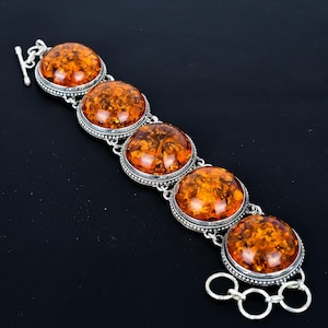 Peut inclure: Un bracelet en argent orné de cinq grandes pierres d'ambre ovales. Les pierres d'ambre sont d'une couleur orange chaude et translucide avec des motifs internes. Le bracelet a un fermoir à bascule et un sertissage argenté décoratif autour de chaque pierre.